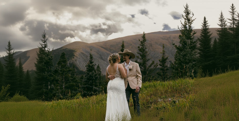 Couple in wedding attire standing in a mountainous landscape with trees and clouds.